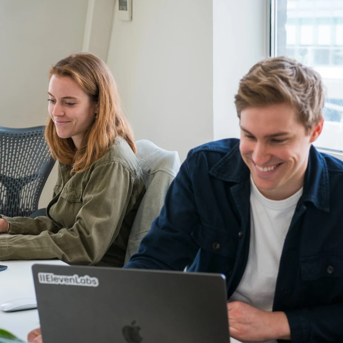 Two people working and smiling at laptops in an office setting.