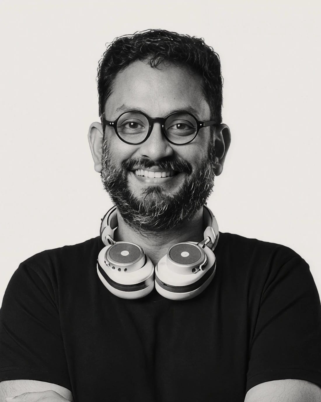 Smiling bearded man wearing round glasses and headphones around his neck.
