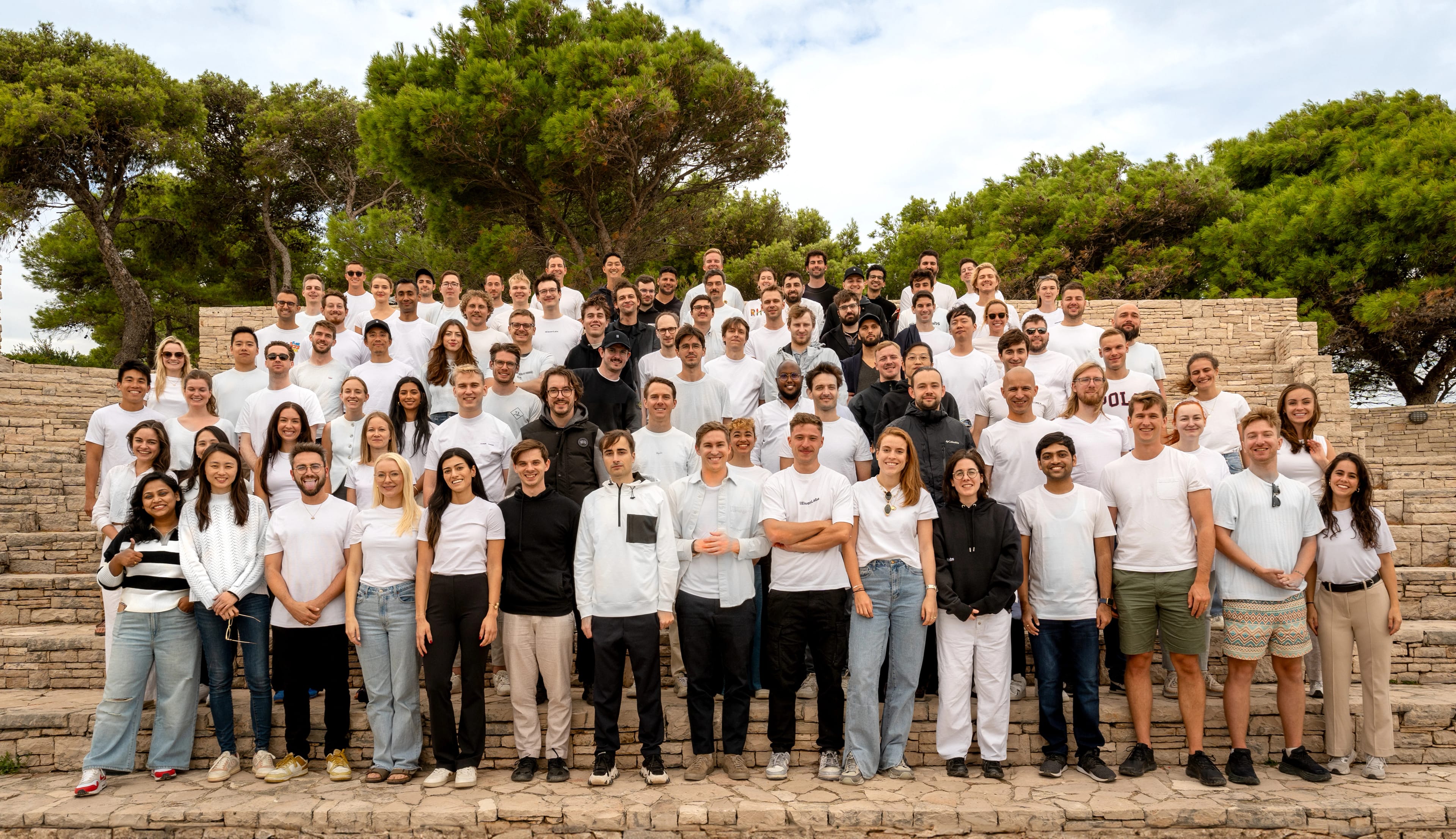 A large group of people posing together outdoors on stone steps, wearing casual white clothing.