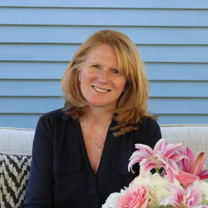 A woman with shoulder-length red hair smiling, sitting outdoors with a blue wooden wall in the background and a bouquet of pink and white flowers in front of her.