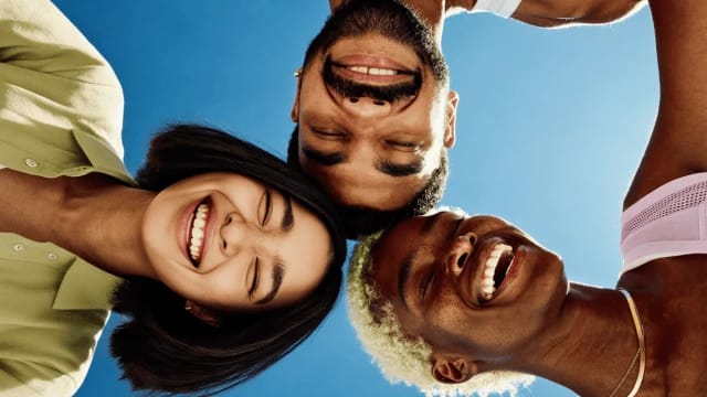 Three friends smile together in a close-up selfie against a bright blue sky.