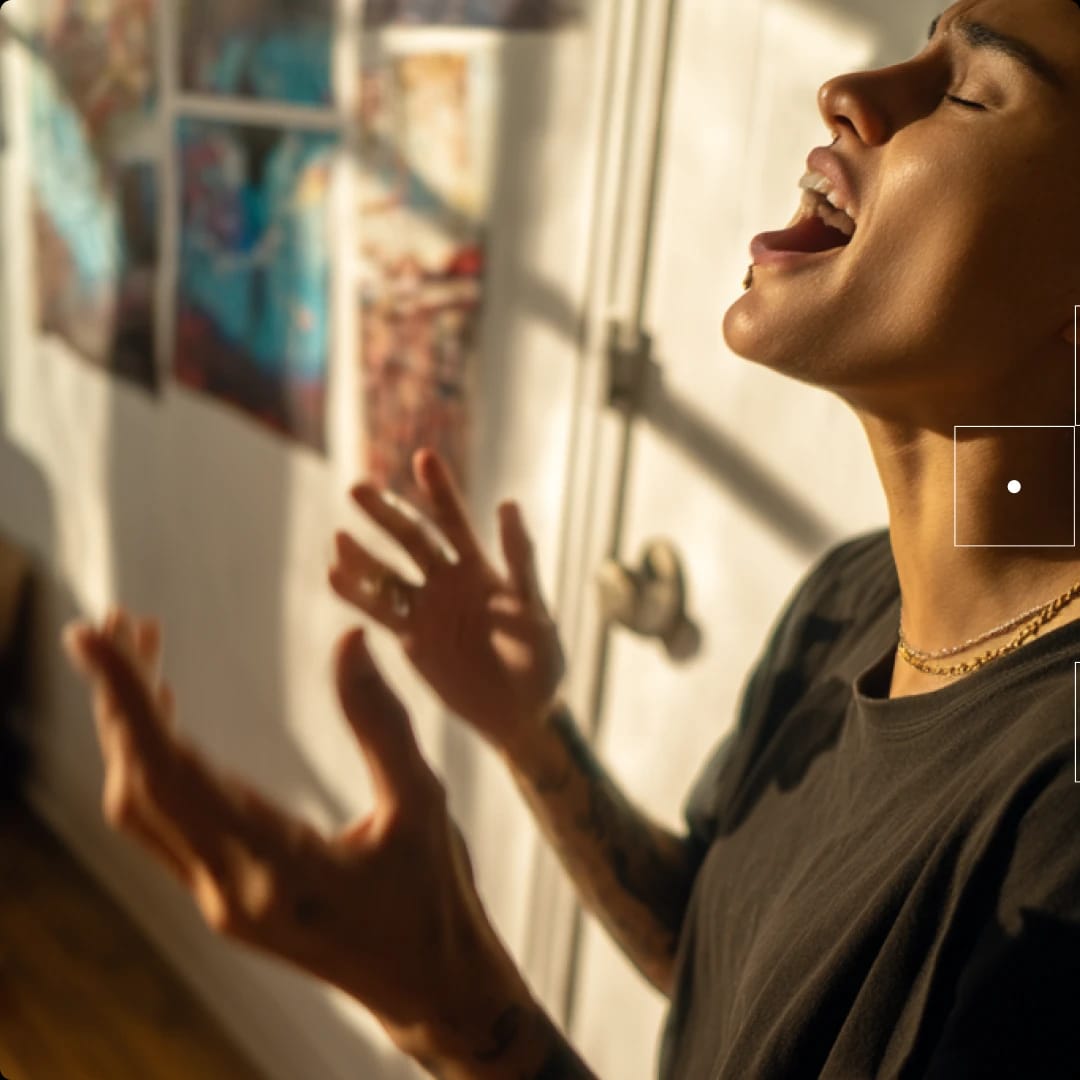 A person singing passionately with eyes closed, standing near a window with sunlight, and artwork on the wall behind.