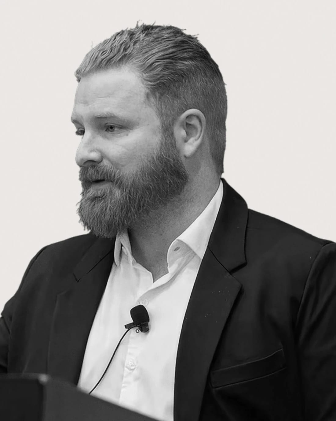 Bearded man in a suit speaking into a lapel microphone.