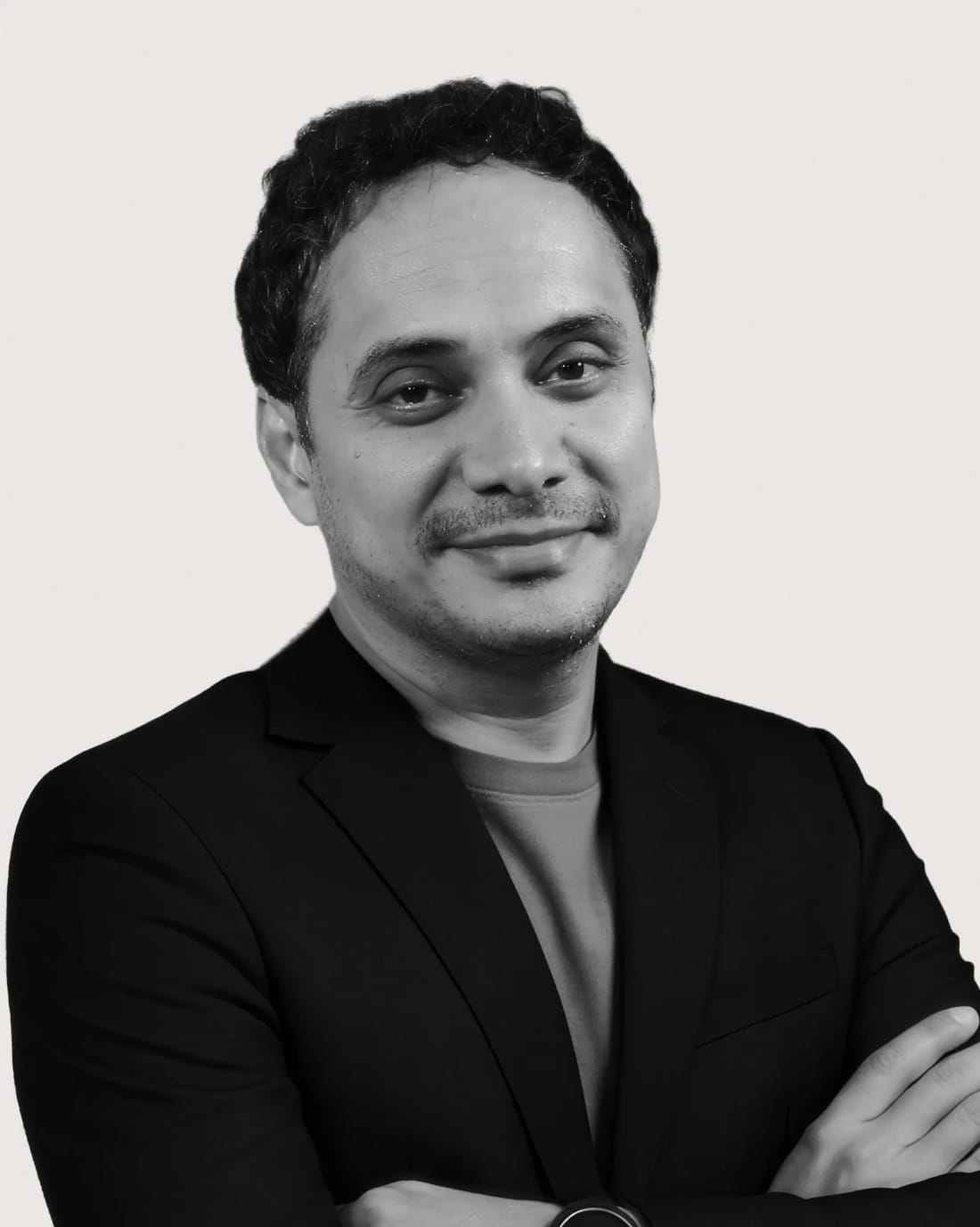Smiling man in a blazer with arms crossed against a plain background.