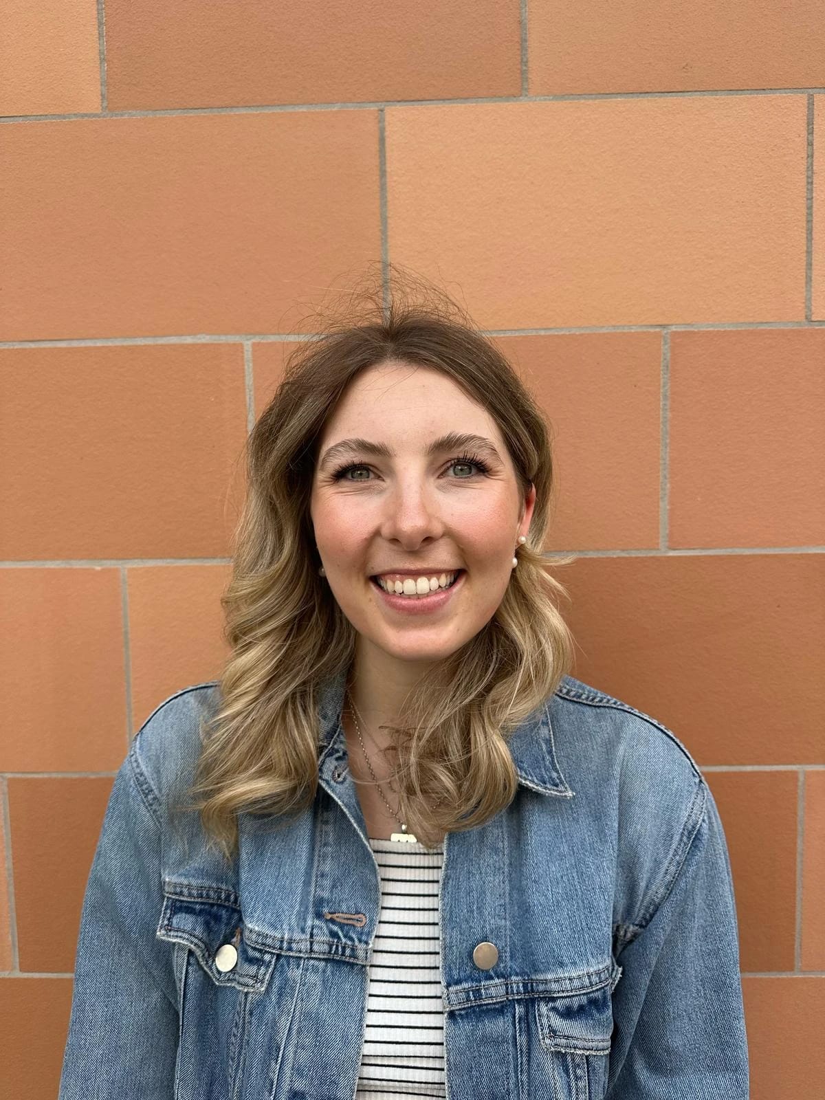 Smiling woman with shoulder-length hair wearing a denim jacket against a brick wall.