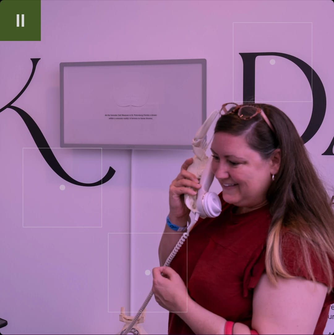 A woman smiling while talking on a vintage telephone with a skull-shaped base, in front of a display with large black letters on a purple wall.