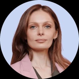 A woman with long, wavy brown hair and fair skin, wearing a pink blazer and a neutral expression.