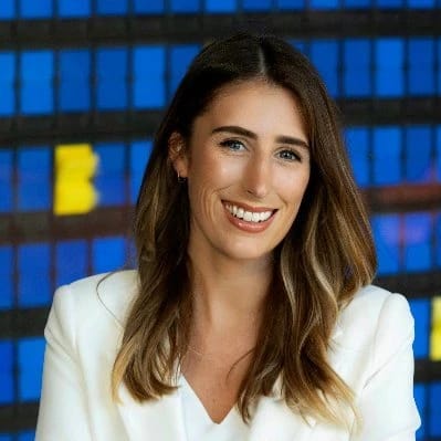 Smiling woman in a white blazer standing in front of a blue stained-glass window.