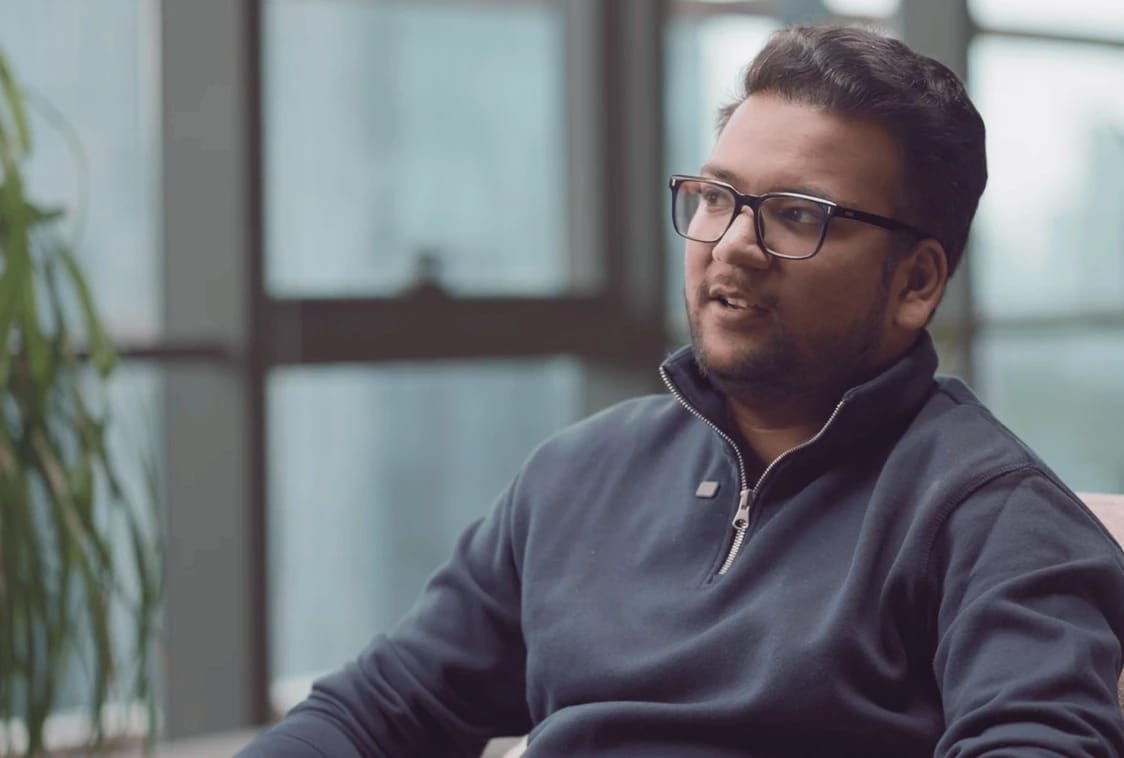 Man wearing glasses and a dark sweater sits indoors, looking to the side.