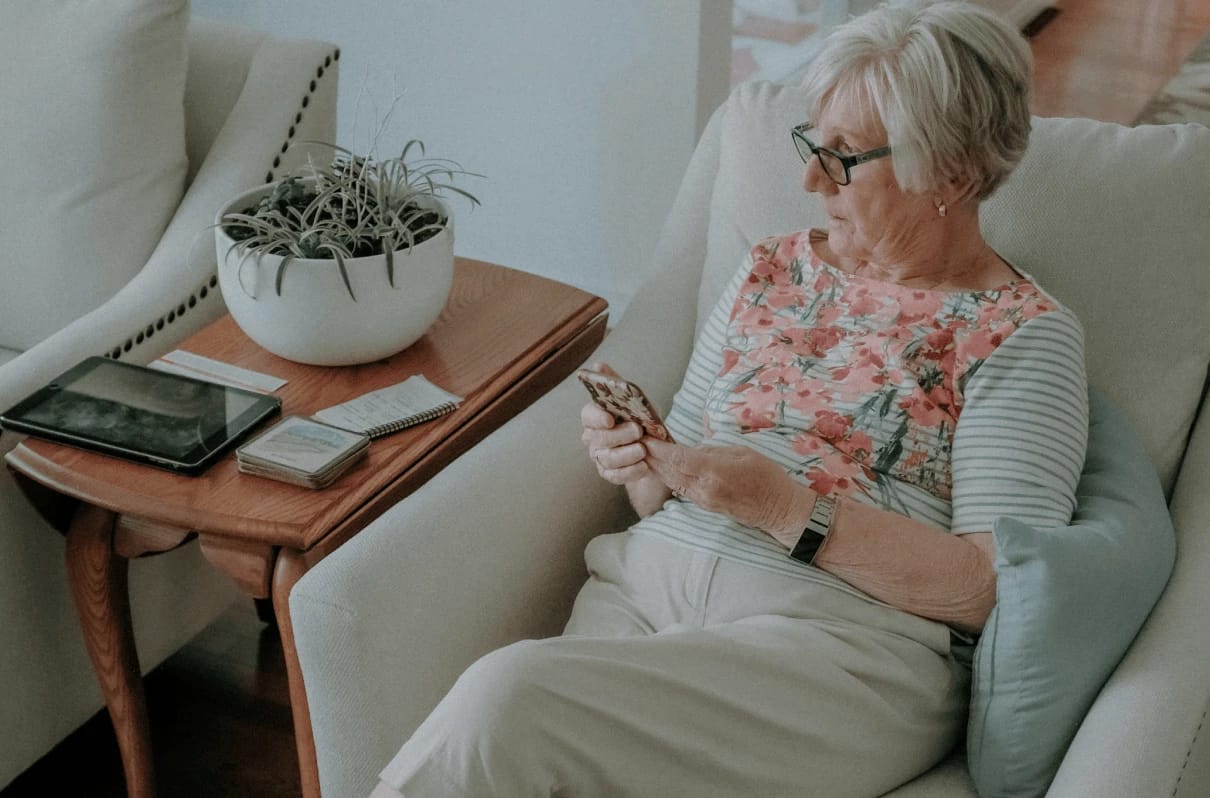 Older woman relaxing in an armchair and looking at her smartphone beside a side table.