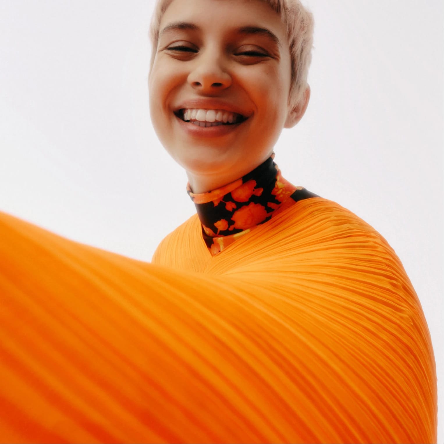 Smiling person in an orange pleated garment and patterned scarf.