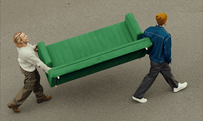 Two people carry a green sofa across a paved street.