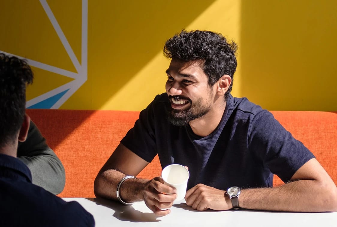 Smiling man holding a paper cup while talking with a companion at a café table.