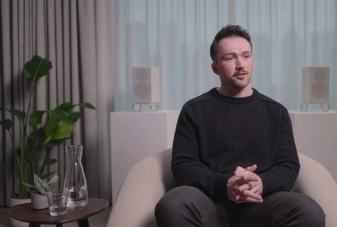 Man in a black sweater sits in an armchair during an interview.