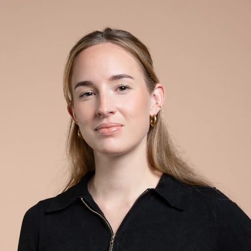 A young woman with long blonde hair, wearing a black zip-up top and gold hoop earrings, posing against a beige background.