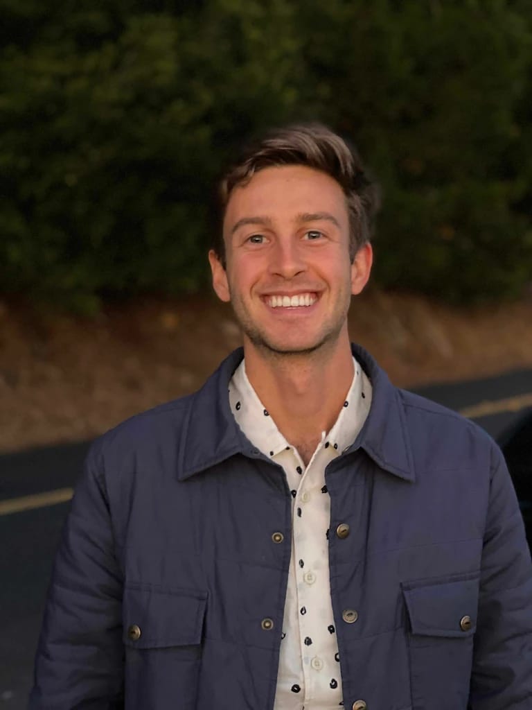 A person smiling, wearing a blue jacket and a patterned shirt, standing outside with greenery in the background.