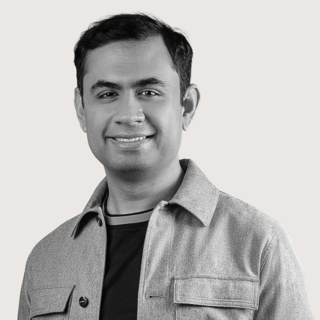 Smiling man wearing a light jacket and dark shirt against a plain background.