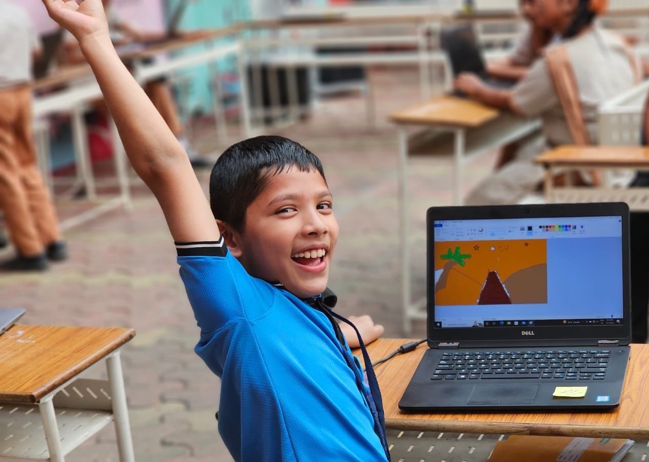 A child in a blue shirt joyfully raises their hand near a laptop displaying digital artwork.