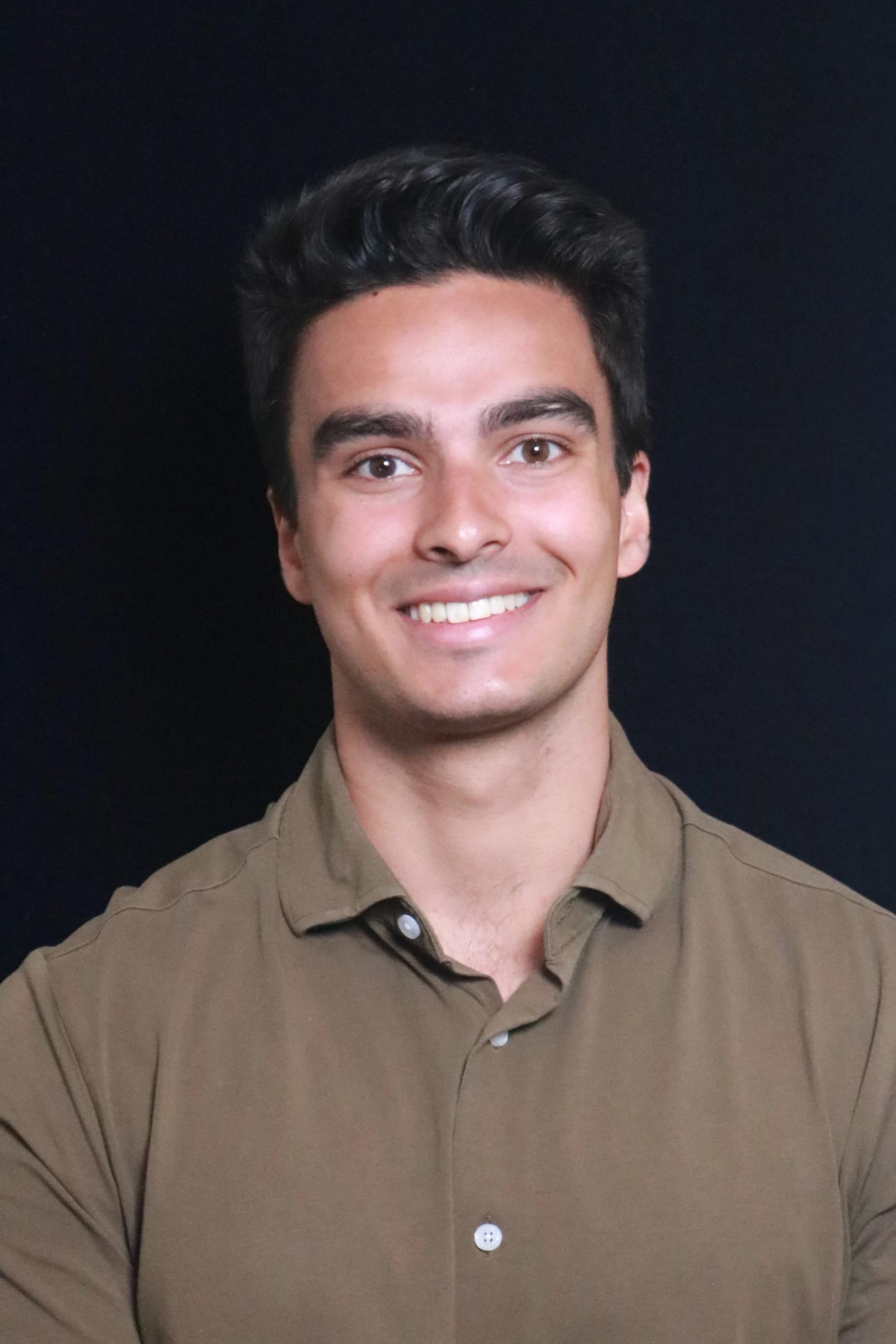 Smiling man with dark hair wearing a brown collared shirt against a dark background.