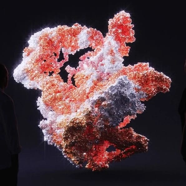 A large, abstract, illuminated sculpture with pink, red, white, and black colors, viewed against a dark background with two people observing it.