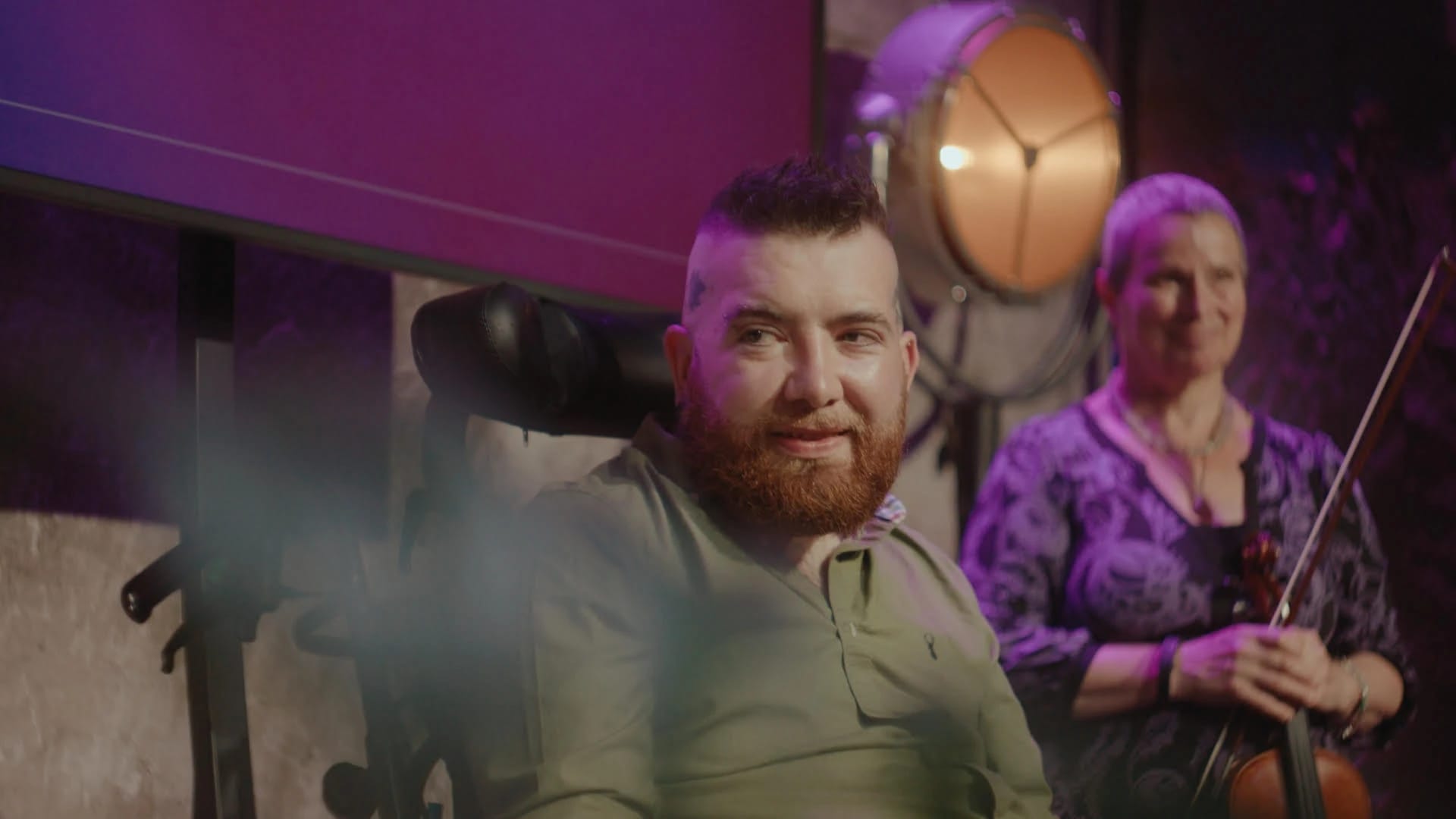 Man with a beanie in a wheelchair smiling next to a woman holding a violin.