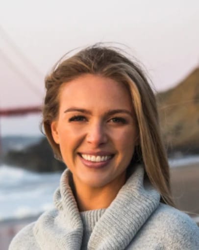 Person smiling on a beach with a bridge and hills in the background.