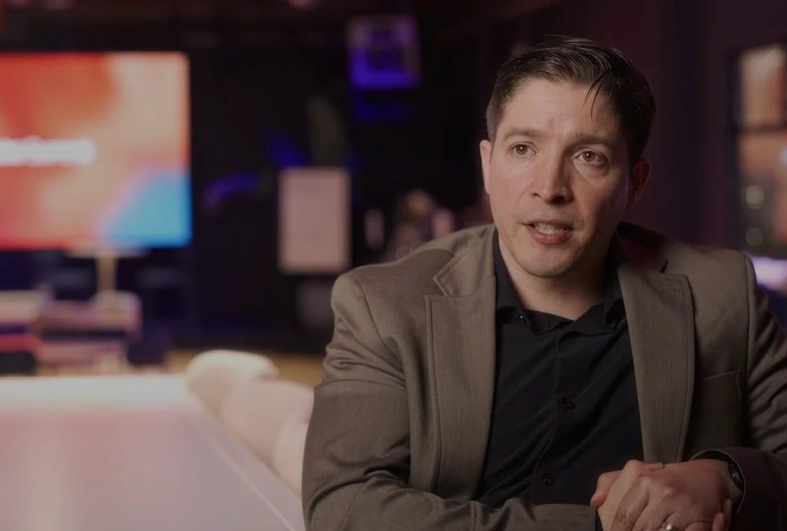 Man in a blazer speaks during an interview in a dimly lit lounge.