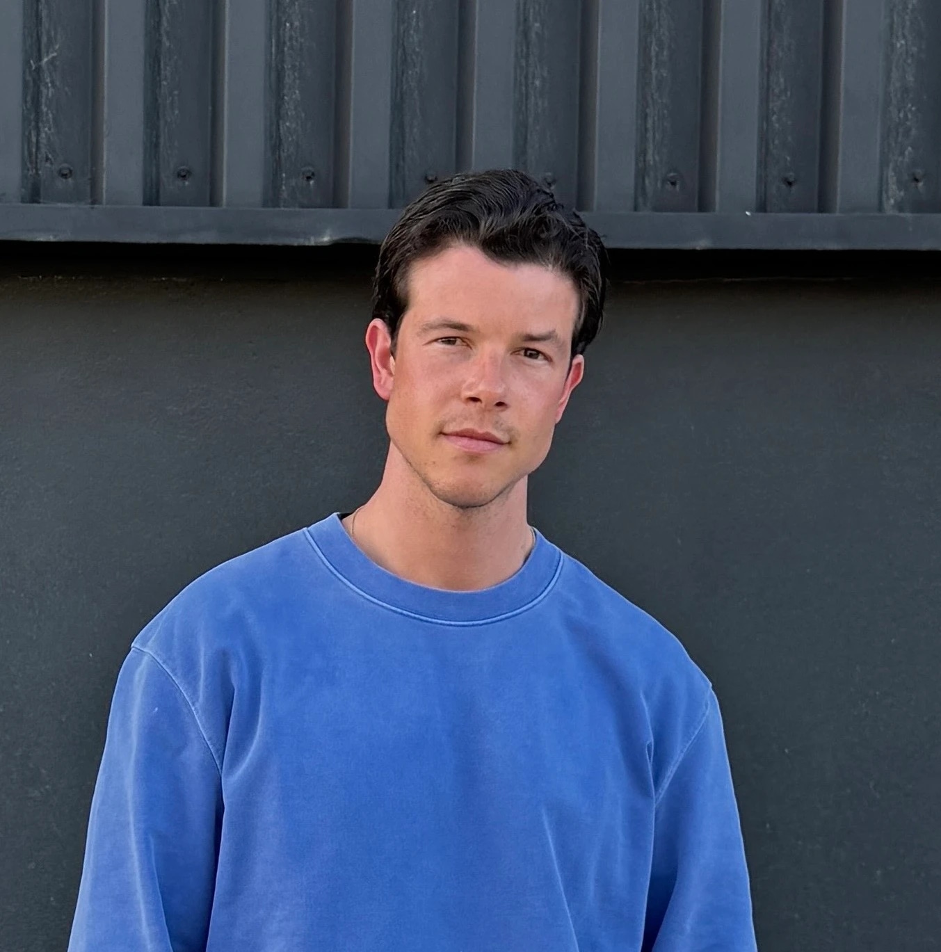 Man wearing a blue sweatshirt standing against a dark wall.