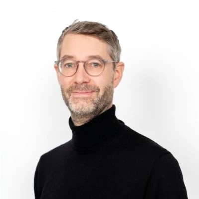 A person with glasses and a black turtleneck, smiling against a white background.