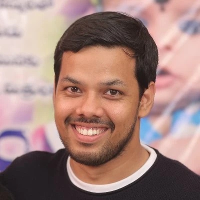 A smiling man with short dark hair and a beard, wearing a black sweater with a white collar.