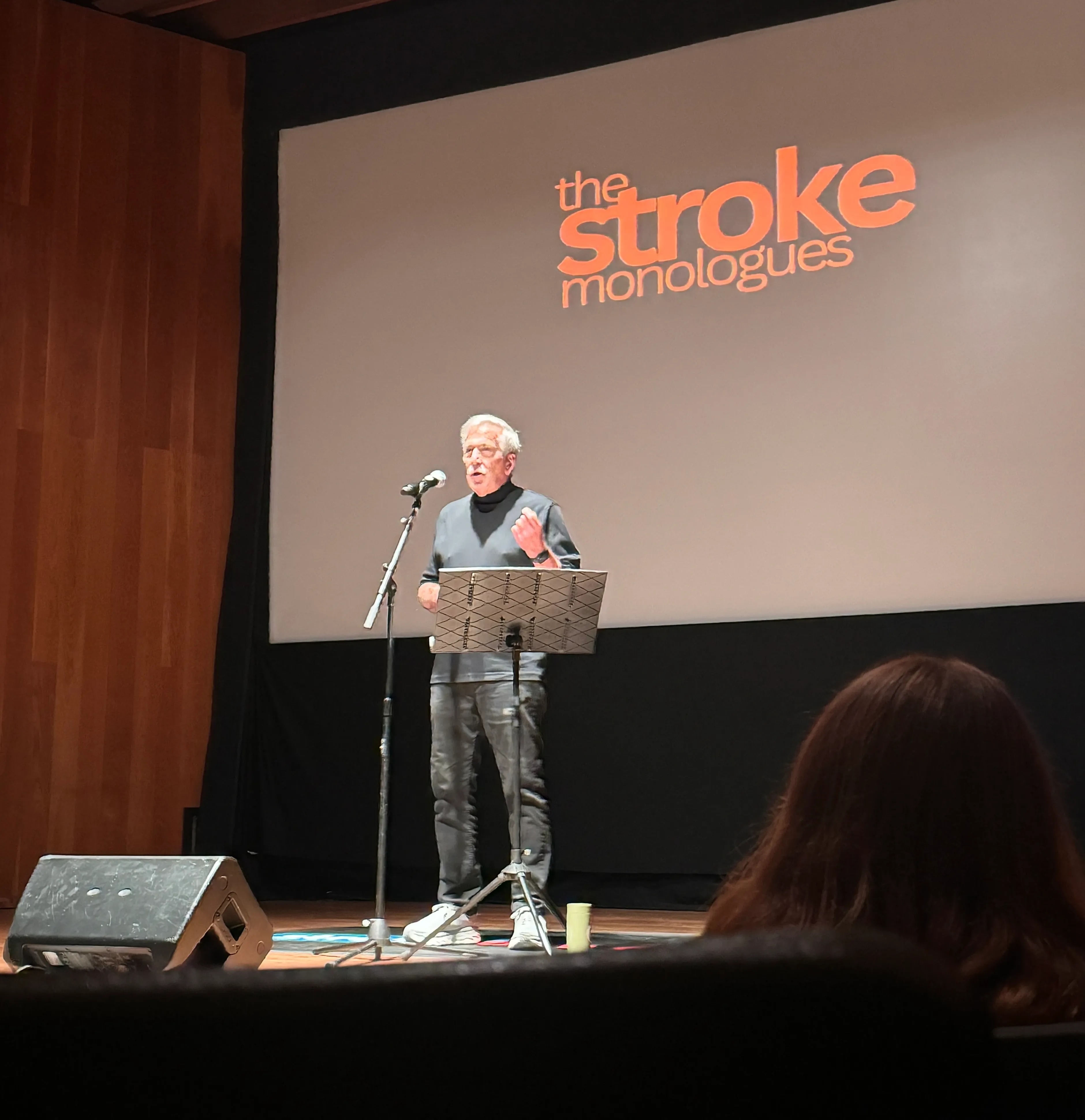 Man standing on stage presenting a speech, behind him a screen says "The Stroke Monologues"