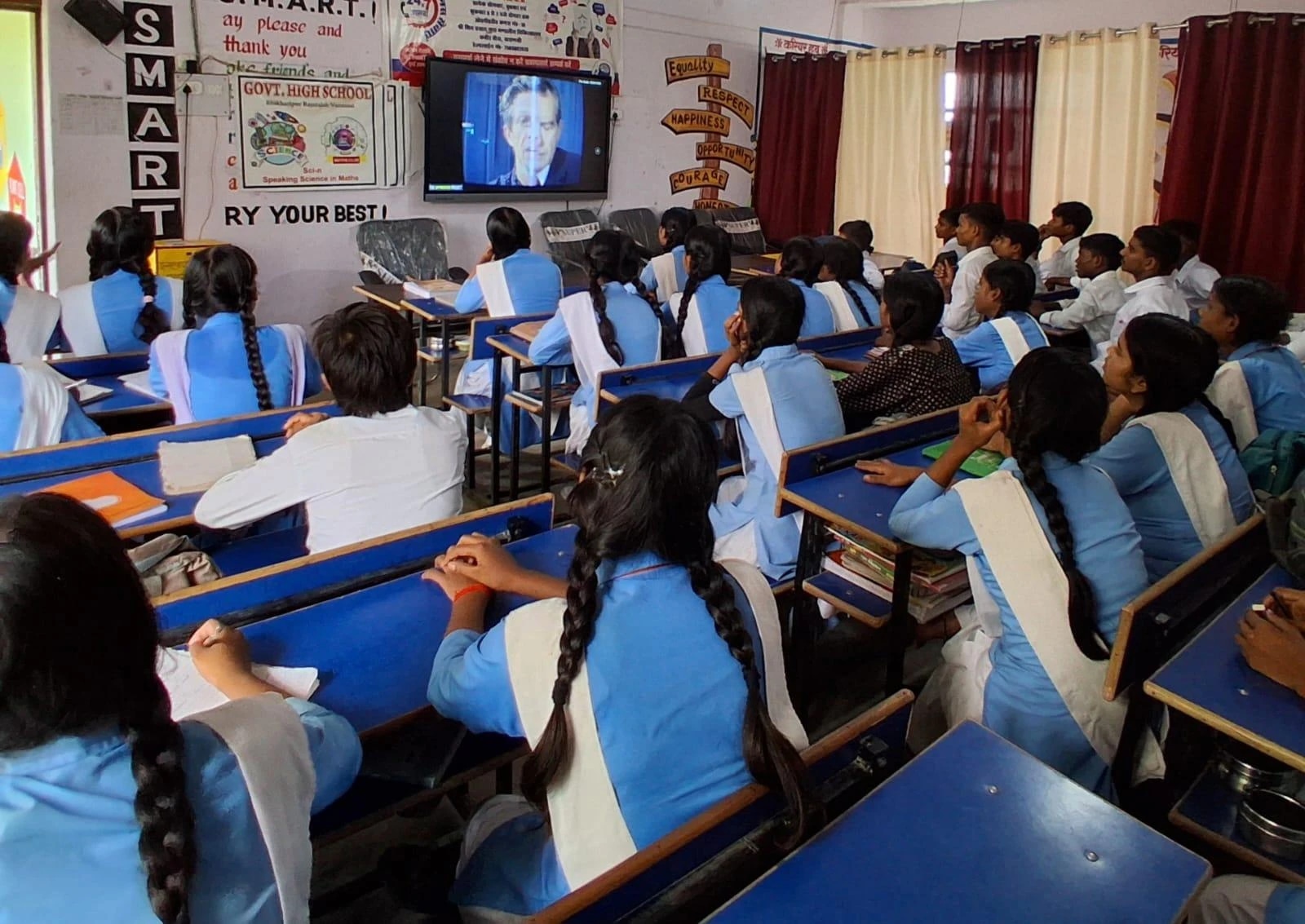 Students in a classroom watching a video lesson on a screen.