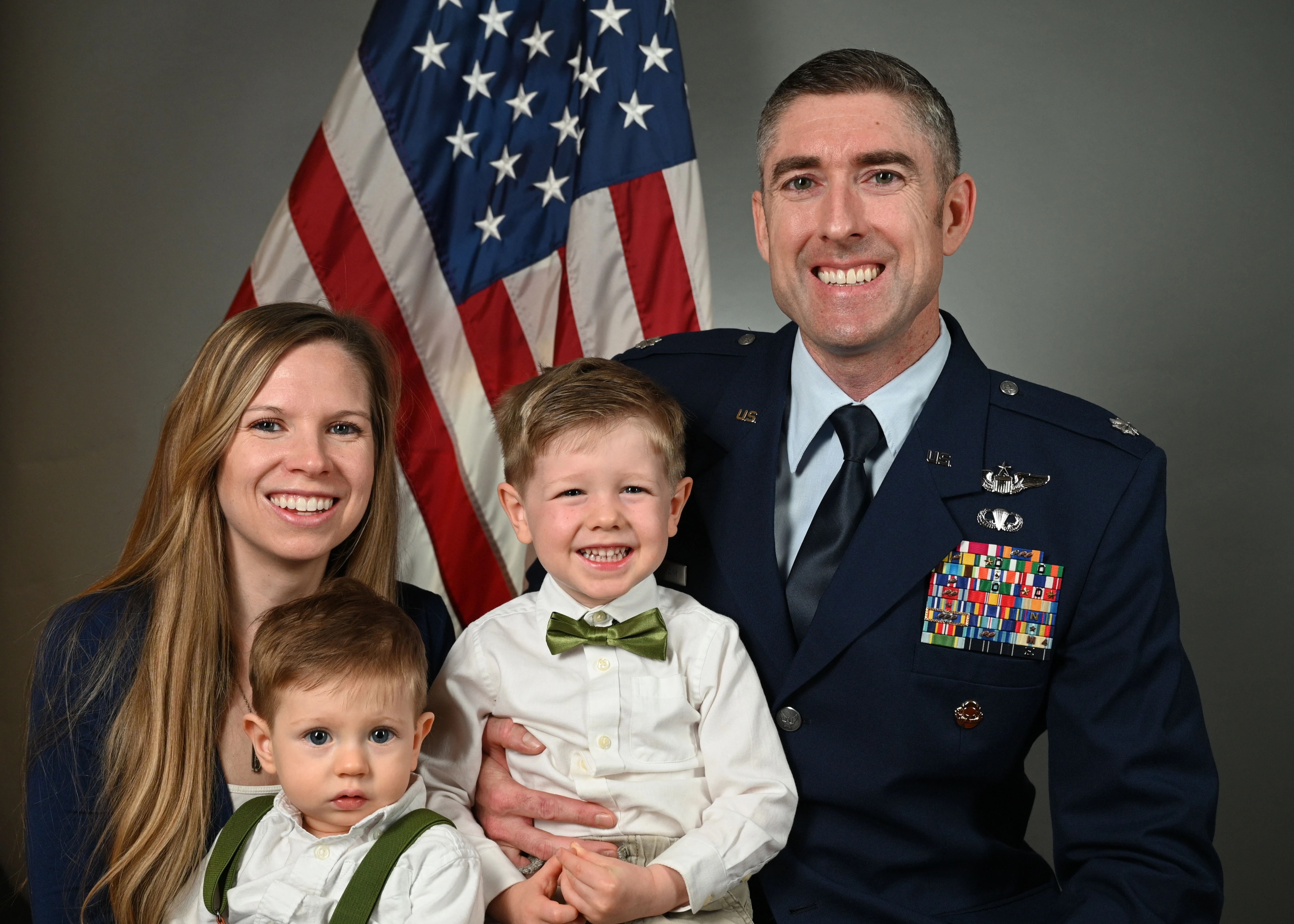 Lt Col Thomas Brittingham poses with his wife, Jessi, and their two young sons in front of the American flag. Thomas is wearing his U.S. Air Force uniform, and the family is smiling.
