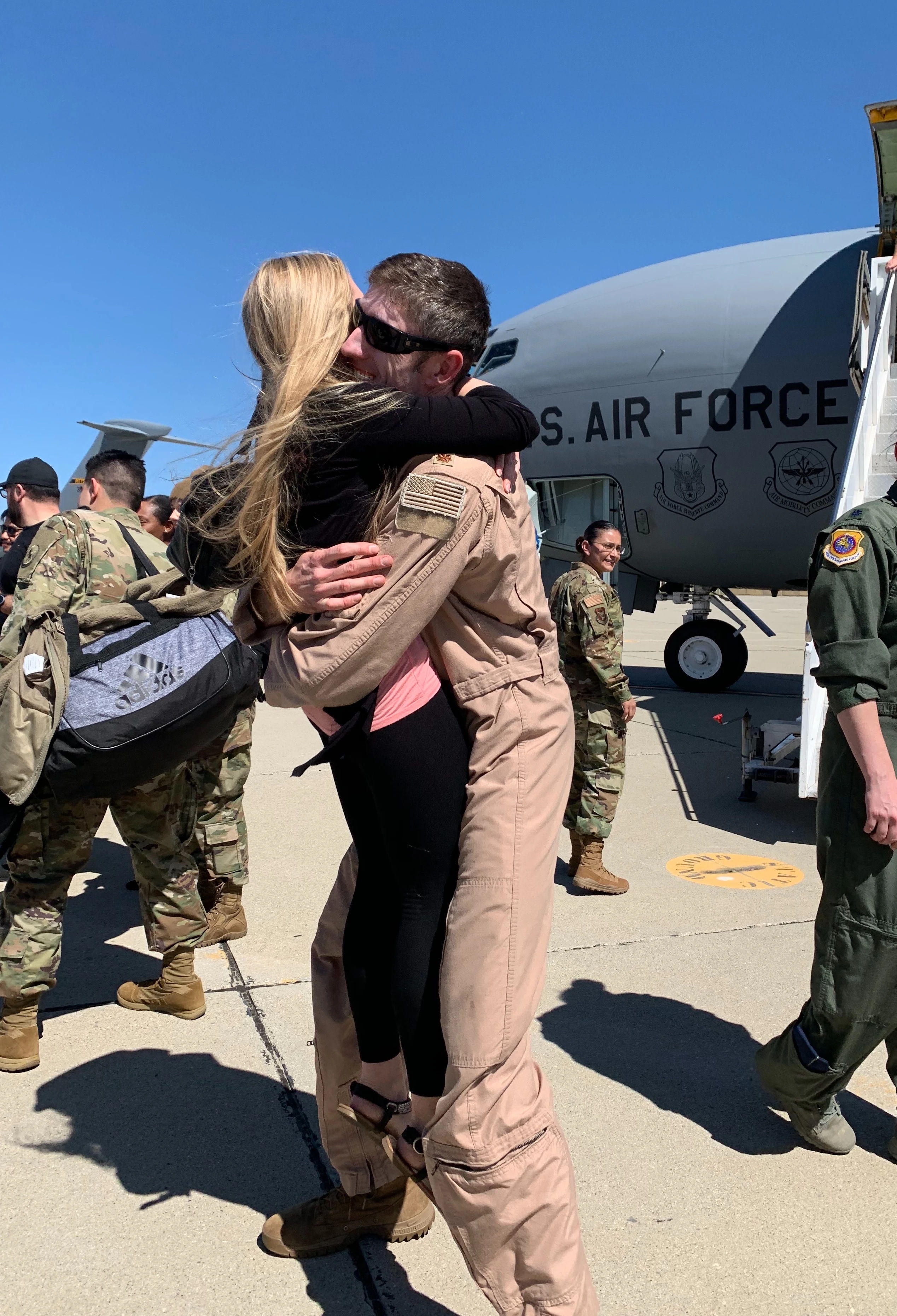 Lt Col Thomas Brittingham embraces his wife, Jessi, on the flight line after returning from deployment. A U.S. Air Force aircraft and service members are visible in the background.