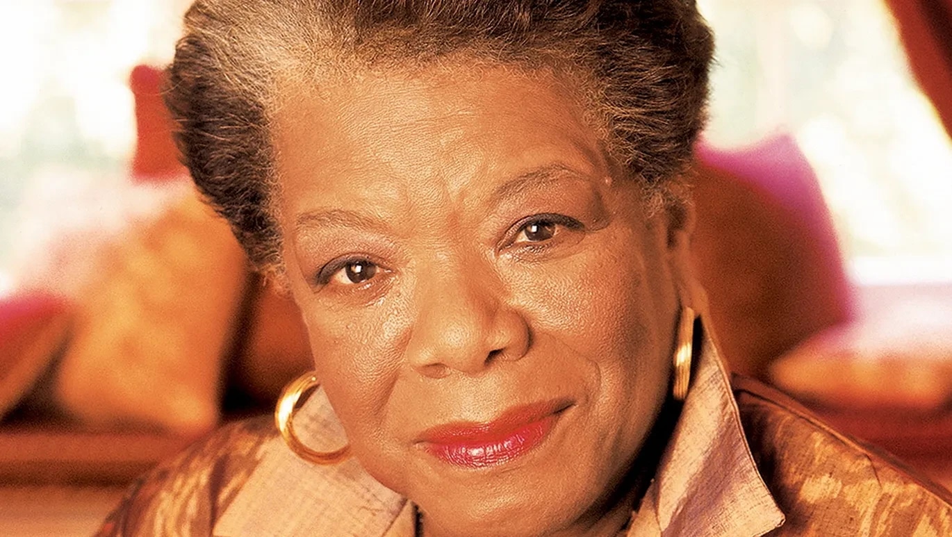 A close-up of an elderly woman with short, styled hair, wearing gold hoop earrings and a patterned blouse, smiling softly.