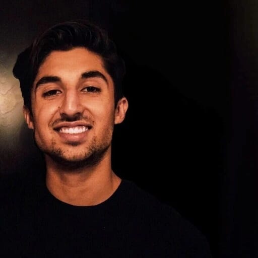 A young man with dark hair and a beard smiling at the camera against a dark background.