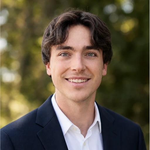 Professional headshot of a smiling man in a navy suit and white shirt outdoors.