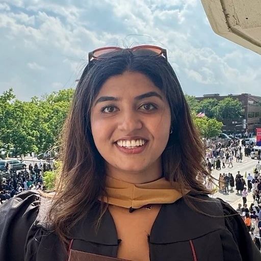 Smiling graduate in academic regalia poses outdoors with a busy crowd behind her.