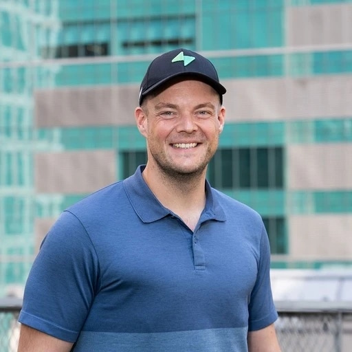 A smiling man in a blue polo shirt and black cap standing outdoors in front of a modern building.