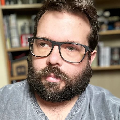 A man with glasses and a beard looking to the side in a room with bookshelves.