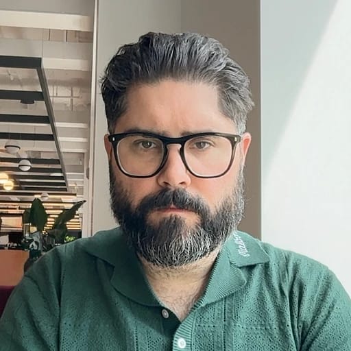 Bearded man wearing glasses and a green shirt, seated indoors.