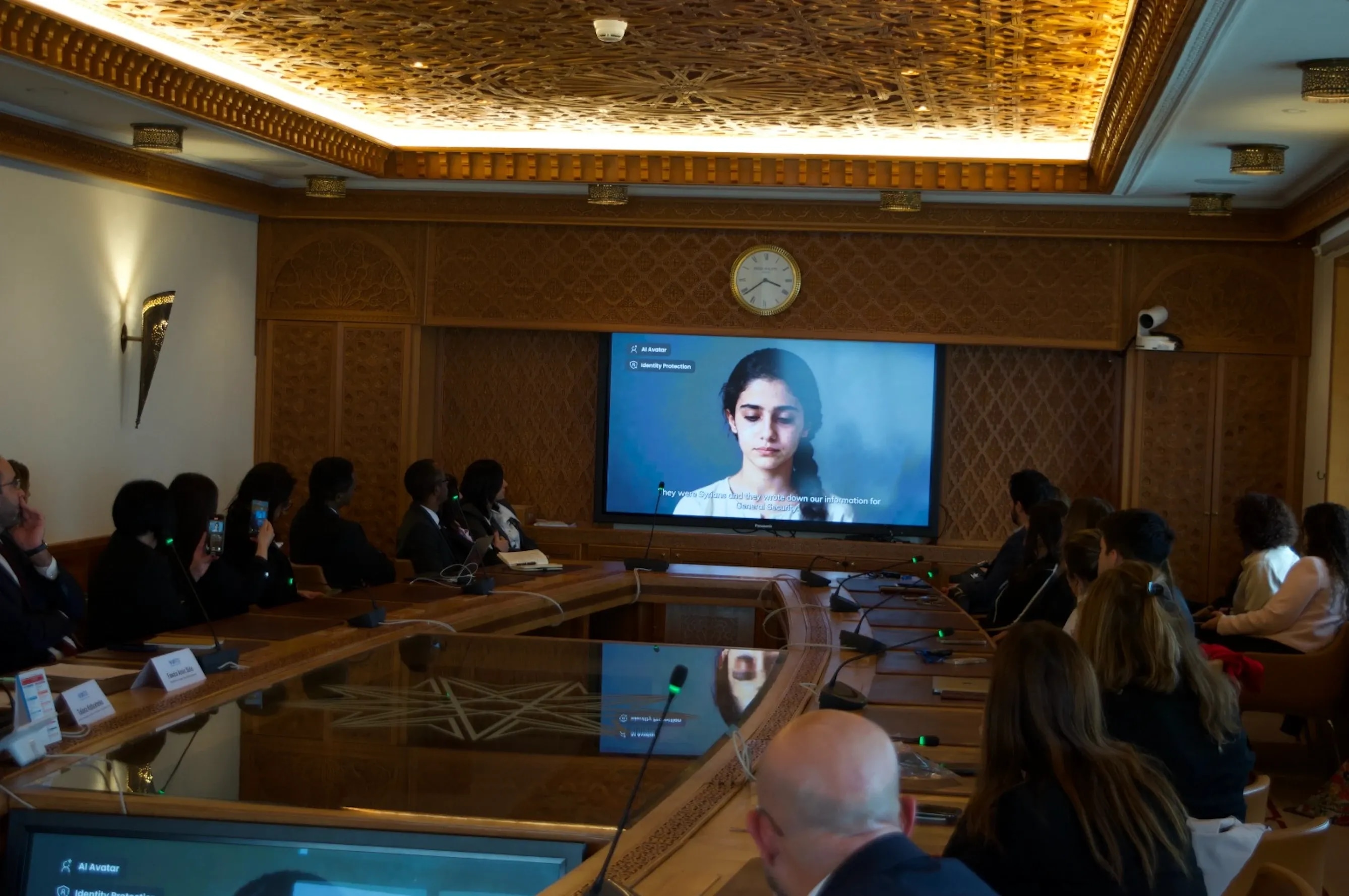 A group of people sit around a large conference table in an ornate meeting room with carved wooden walls and ceiling details. They are watching a large screen at the front of the room displaying a video of a woman with dark hair speaking.