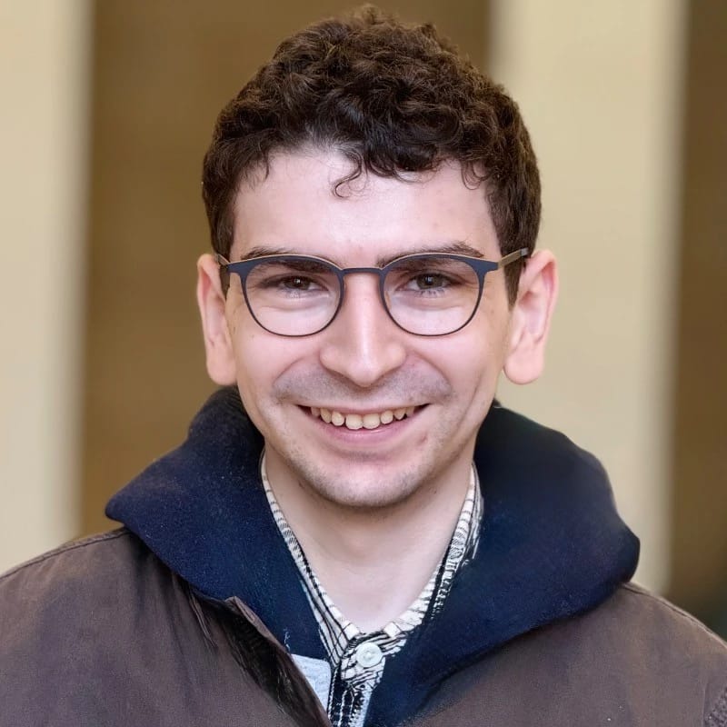 Picture of a man smiling with a blue hoodie and brown shirt.