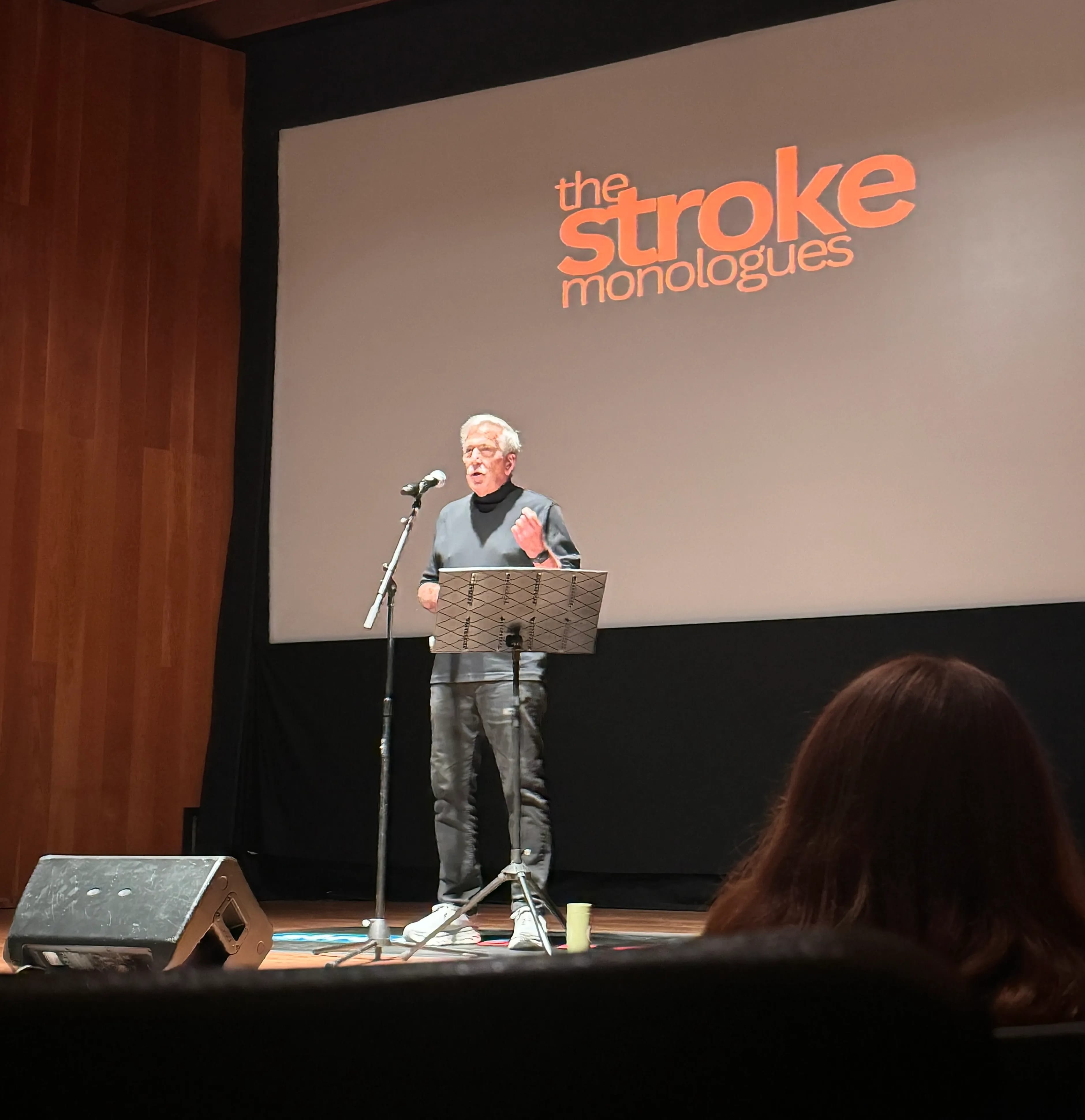 Man standing on stage presenting a speech, behind him a screen says "The Stroke Monologues"