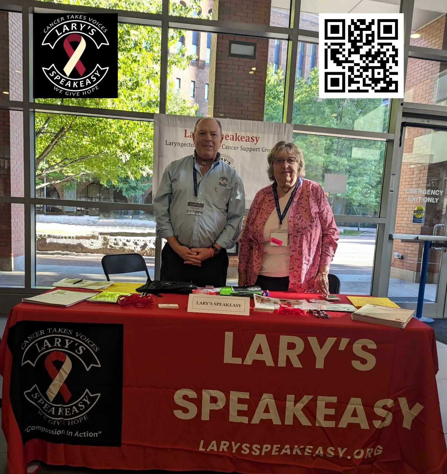 Two Board of Directors members from Lary's Speakeasy, Stephen Cooper and Brooke Elkan-Moore, standing behind a red table display at Thomas Jefferson University Hospital in Philadelphia. The table features the organization's logo with their slogan "Cancer Takes Voices, We Give Hope" and website information. Behind them is a banner and a QR code, with large windows showing a tree-lined street outside.