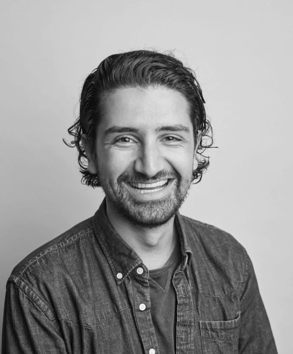 A smiling man with wavy hair and a beard, wearing a denim shirt, in black and white.