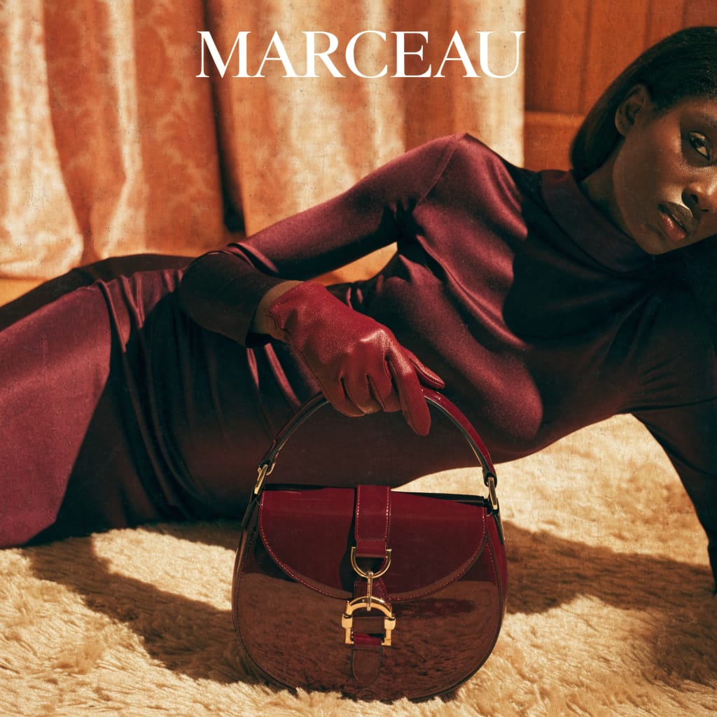 This is a static, high-fashion advertisement with a warm, luxurious, and slightly dramatic mood. The setting appears to be an indoor studio with soft, diffused lighting, creating distinct shadows. The visual style is realistic, emphasizing the textures and sheen of the garments and accessories. The central subject is a dark-skinned woman, elegantly posed in a reclining position, dressed in a form-fitting, dark burgundy dress and matching leather gloves. She holds a glossy, deep red handbag with gold hardware, which is the product being advertised, bearing the brand name "MARCEAU" prominently at the top. The color palette is dominated by rich, warm tones of burgundy, dark red, and golden-orange, with high saturation that creates a sophisticated and alluring aesthetic. The background features a textured, light orange curtain, and the woman is lying on a plush, light brown or beige rug, adding to the opulent feel.