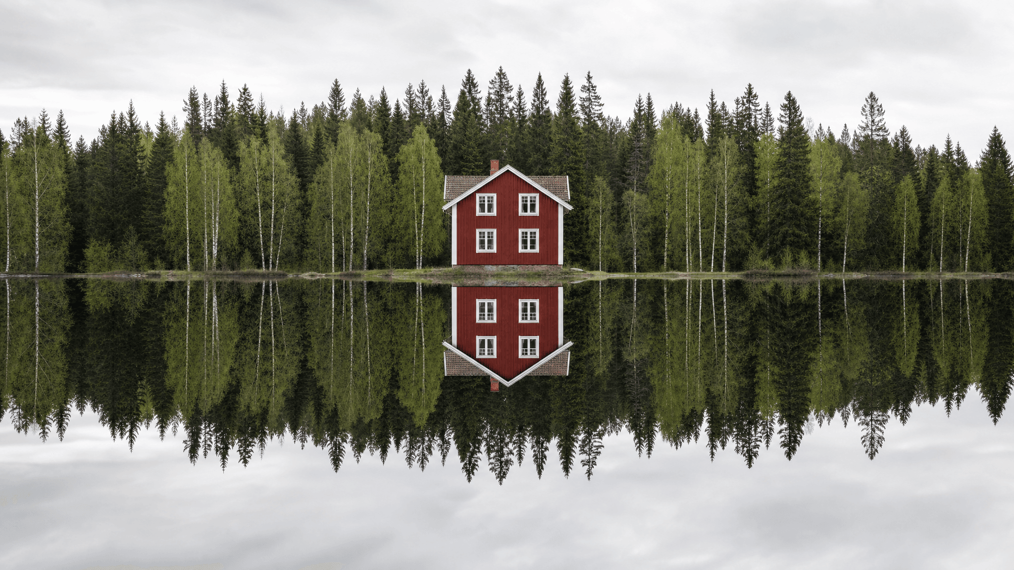 This video presents a serene, static shot of a vibrant red cabin nestled at the edge of a lush green forest, perfectly reflected in the calm, dark water of a lake. The overall mood is peaceful and tranquil, with a slightly mysterious or isolated emotional tone. The setting is an outdoor, natural environment, likely in a Nordic or northern European landscape, emphasizing nature and solitude. The visual style is realistic and artistic, highlighting symmetry and contrast. The color characteristics feature cool tones in the sky and water, balanced by the warm, striking red of the house and the various shades of green in the dense forest. The color palette prominently includes red, white, dark green, light green, and muted grays, creating a captivating and balanced composition.