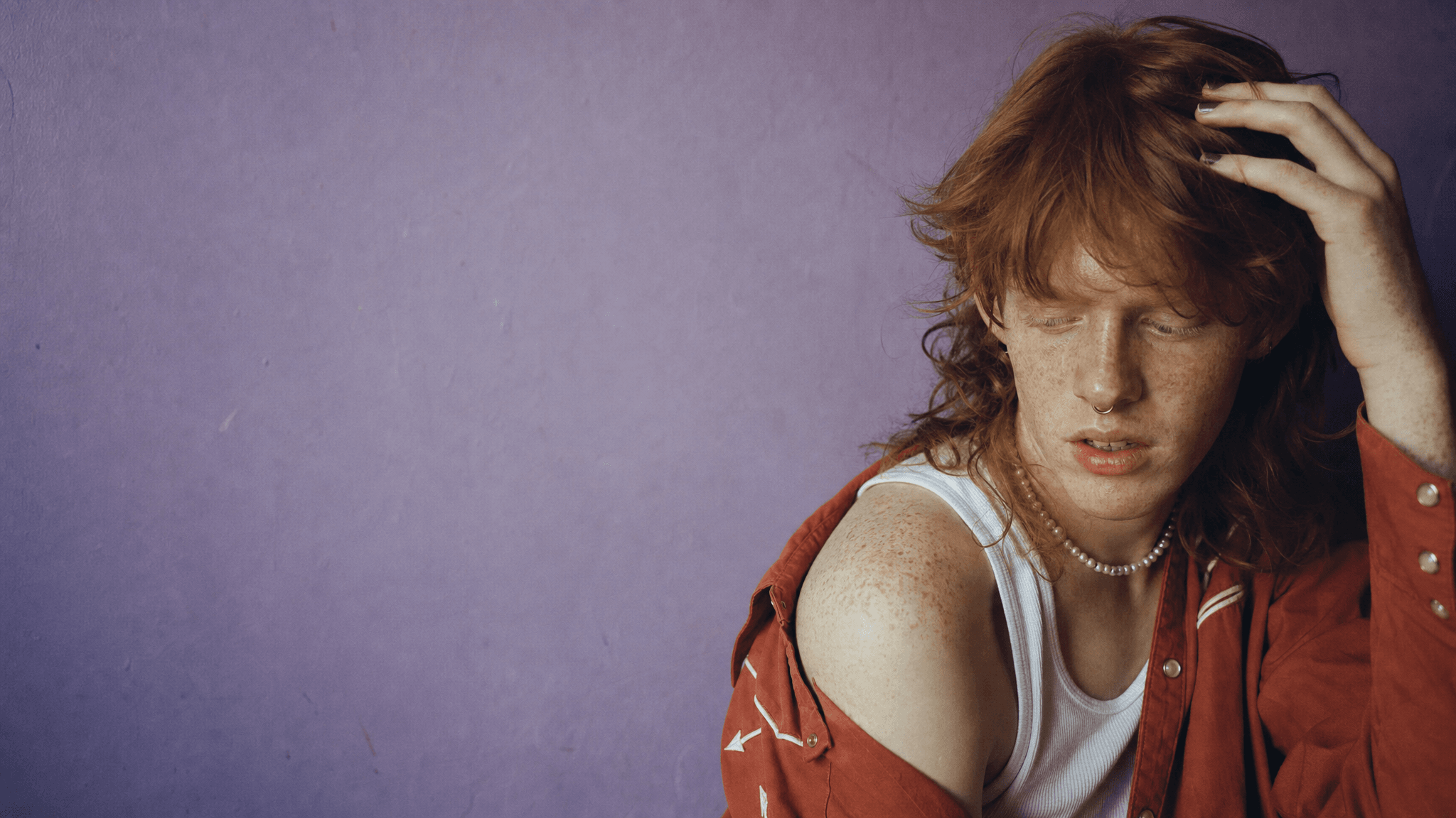 This video features a close-up, static shot of a young person with vibrant red hair and numerous freckles. The mood is calm and contemplative, with the subject's eyes closed and one hand gently touching their hair, revealing green nail polish. The setting is indoors, against a plain, light purple wall. The visual style is realistic and intimate, emphasizing the subject's natural features and a subtle, melancholic emotional tone. The person wears a white tank top, a pearl necklace, and an unbuttoned red shirt with white arrow patterns. The color characteristics include warm skin tones and hair contrasting with the cool purple background, with high saturation creating a vivid, artistic aesthetic. The color palette is dominated by reddish-orange, white, and a soft lavender.