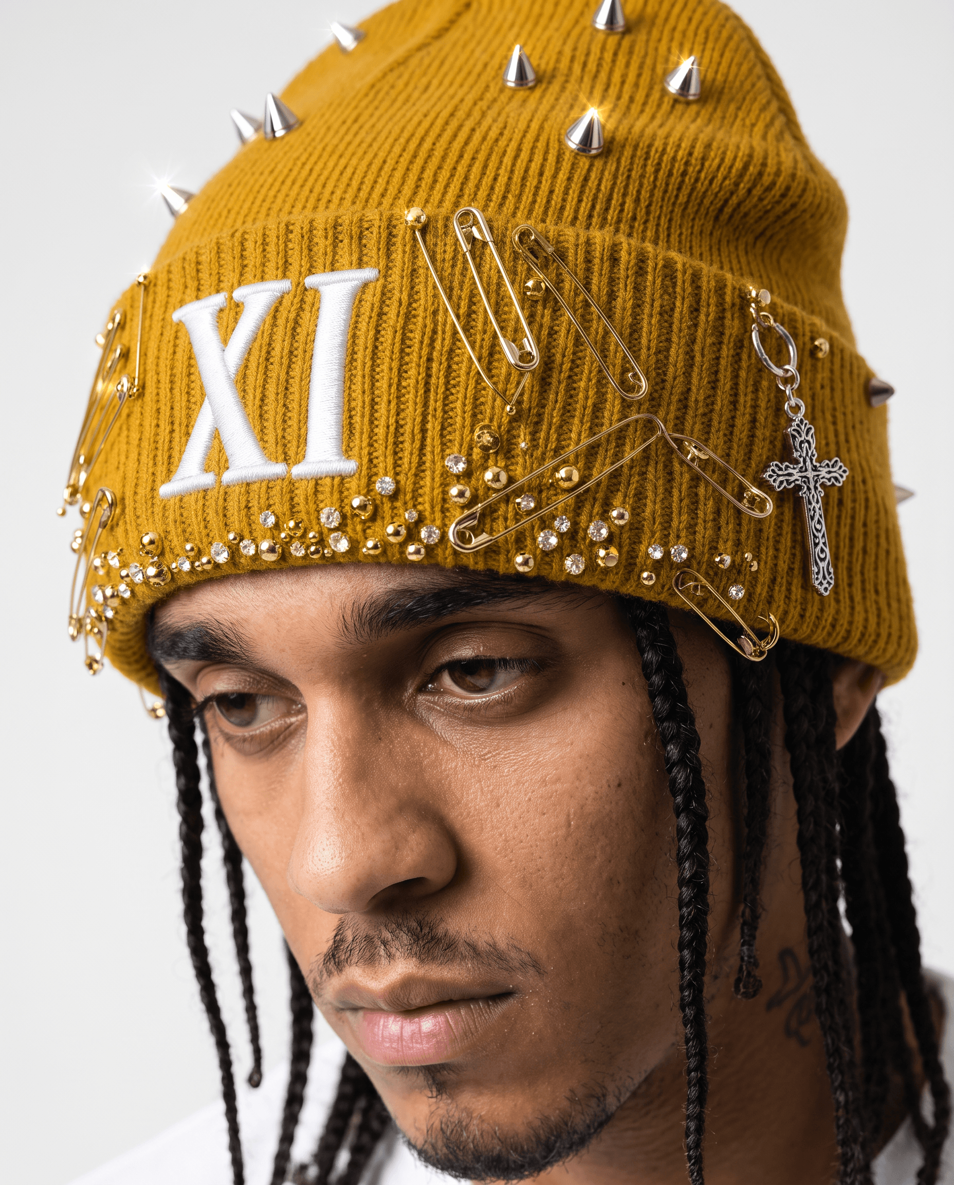This video features a close-up portrait of a young man with dark braided hair, wearing a mustard yellow beanie adorned with metallic spikes, golden safety pins, small gold beads, and rhinestones. A silver cross charm hangs from the side of the beanie. The mood is calm and contemplative, with the man looking downwards, suggesting introspection or a subtle gaze. The setting is a brightly lit indoor studio with a plain white background, emphasizing the subject and his unique headwear. The visual style is realistic and fashion-focused, highlighting the intricate details of the beanie. The color palette primarily consists of warm mustard yellow, cool silver, and gleaming gold against the man's warm skin tones and dark hair, creating a striking contrast.